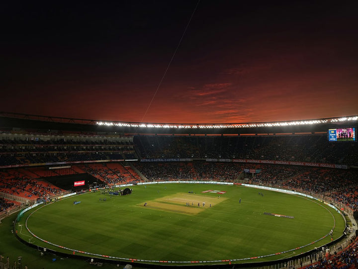 A cricket stadium with a green pitch under a dark red evening sky — illustrative stock photo